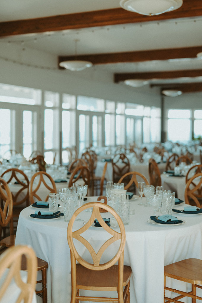 Wedding reception set up with white linens, and blue napkins, and light oak chairs 